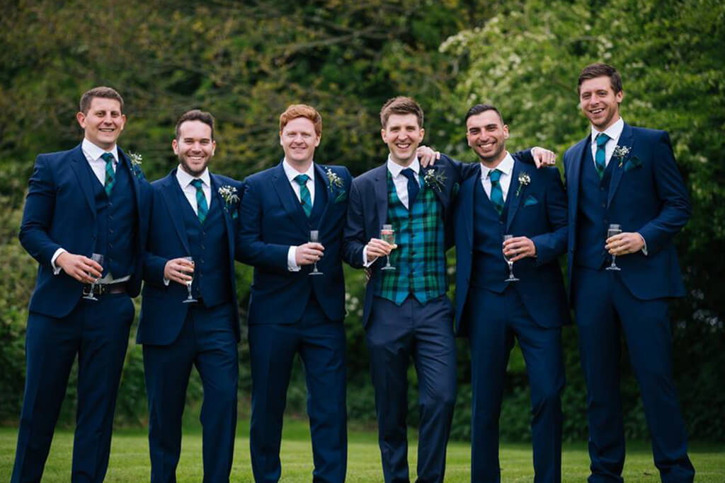 The groom stands with his groomsmen each wearing striking blue suits with tartan waistcoats 