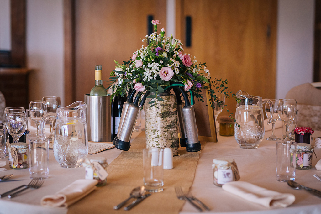 The couple chose quirky table decorations with dairy farming accessories and vases of pretty flowers as table centrepieces at Sandhole Oak Barn