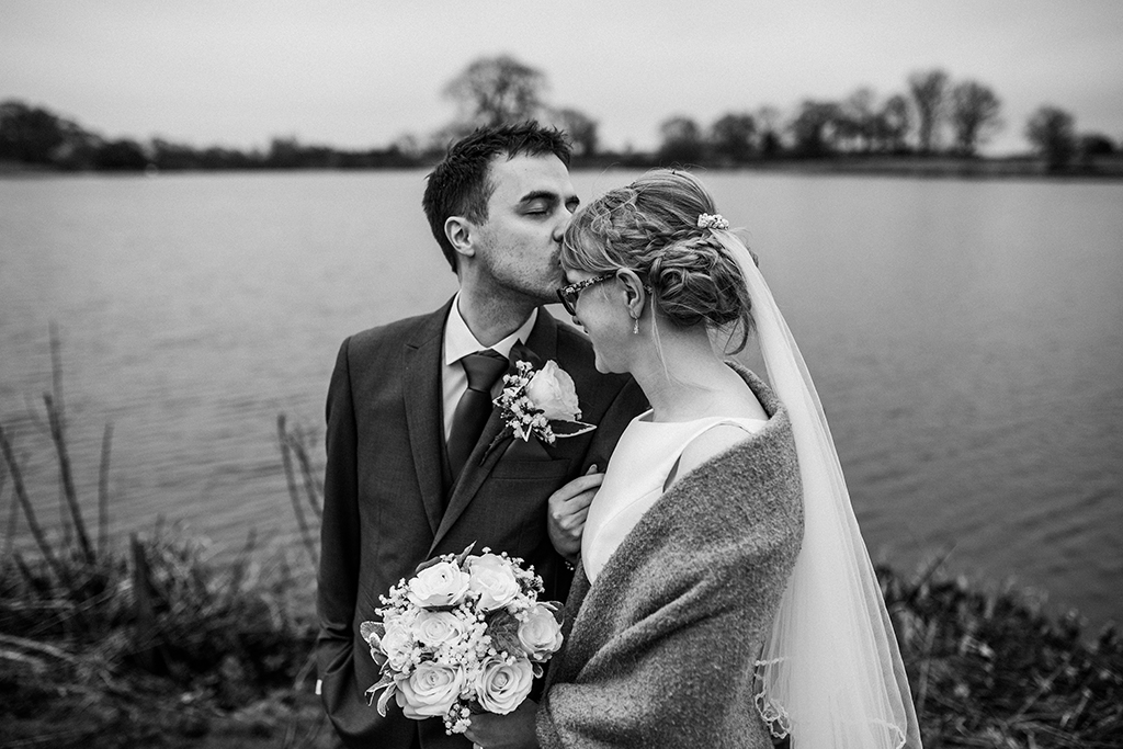 The groom plants a sweet kiss on his wife’s head as they pose for wedding photos at the lake at Sandhole Oak Barn 
