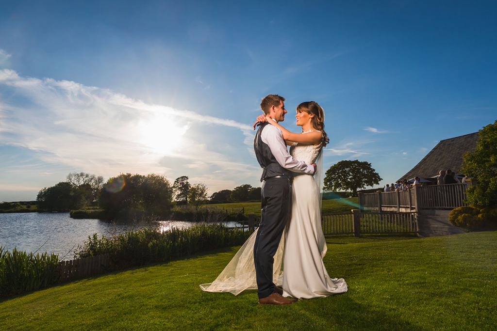 The happy couple pose for wedding photo in front of the lake at Sandhole Oak Barn