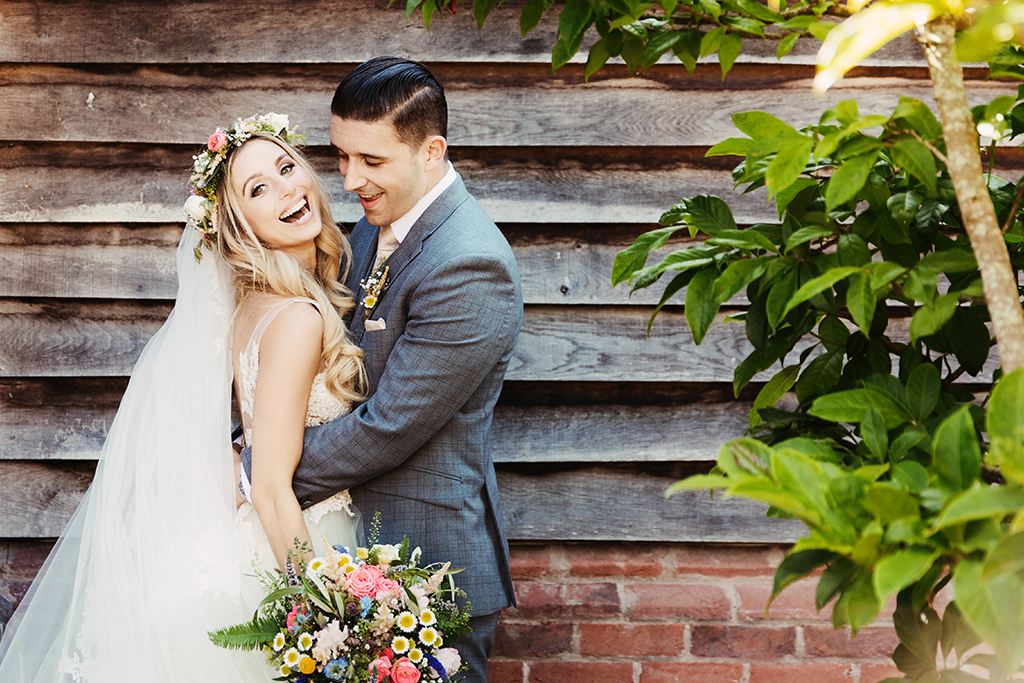 The happy bride wears a pretty floral crown and holds a coordinated bouquet as she poses for a photo with the groom in front of the barn