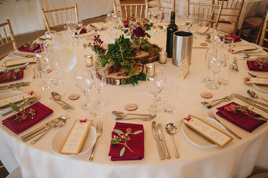 Round wedding tables were decorated with wood slices and pretty flowers with coordinating burgundy napkins at this rustic winter wedding at Sandhole Oak Barn