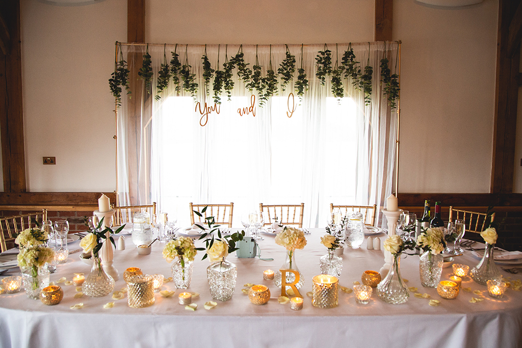 The top table was decorated with pretty vases of cream roses and gold tea light holders and a gold frame with hanging foliage was used as a backdrop at this spring wedding at Sandhole Oak Barn