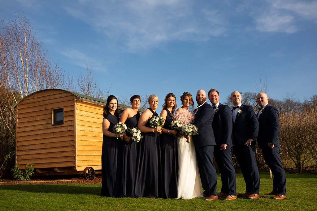 The wedding party pose for a photo by the groom’s hut preparation room at Sandhole Oak Barn 