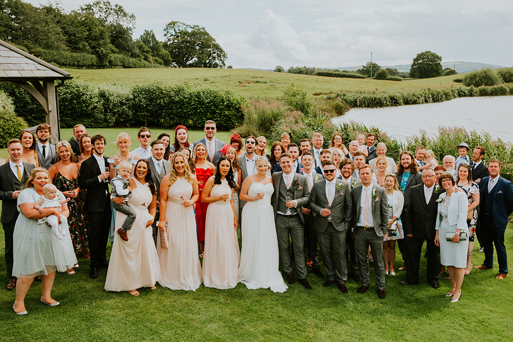 The bride and groom stand together with all their wedding guests on the lawns next to the lake
